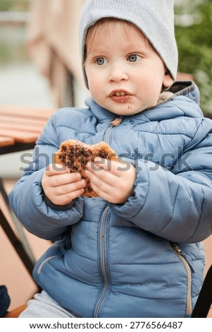 Toddler boy eating cookie while sitting on chair outdoors. Wearing warm jacket and hat on terrace in autumn