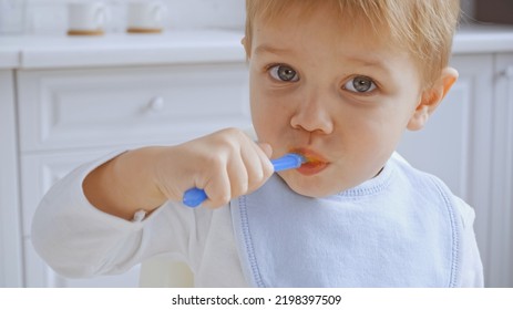 Toddler Boy With Grey Eyes Holding Spoon And Eating Carrot Puree