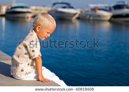 Toddler boy sitting on the pier and looking at the yachts.