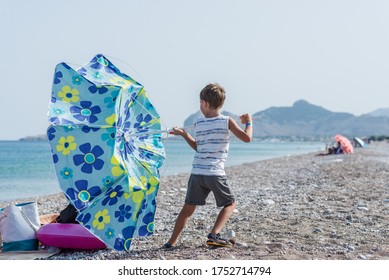 Toddler Boy Standing On Beautiful Pebble Beach Holding Tight To Sun Umbrella As The Wind In Turning It Over.