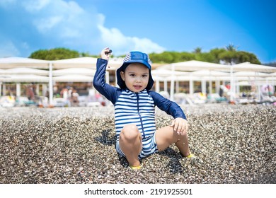 Toddler Boy In A Swimming Suit Plays With Pebbles On The Seashore In Hotel