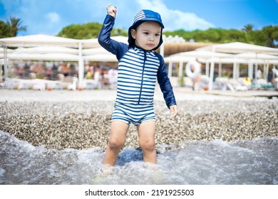 Toddler Boy In A Swimming Suit Plays With Pebbles On The Seashore In Hotel