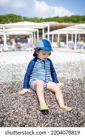 Toddler Boy In A Swimming Suit Plays With Pebbles On The Seashore In Hotel