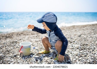 Toddler Boy In A Swimming Suit Plays With Pebbles On The Seashore In Hotel