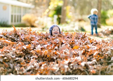 A toddler boy wearing a hat laughs with his eyes closed after jumping into a big pile of leaves. 