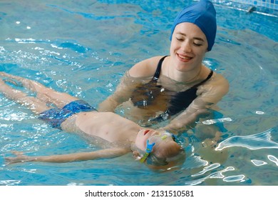 Toddler Child Learning To Swim In Indoor Swimming Pool With Teacher. Floating In The Water, Balancing And General Physical Activity For Kids, Early Development. Boy Kid Trained To Kick Legs And Float