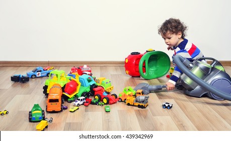 Toddler Cleaning The Messy Room Full Of Toys With A Vacuum Cleaner. Lots Of Toys, Many Cars Piled On The Floor