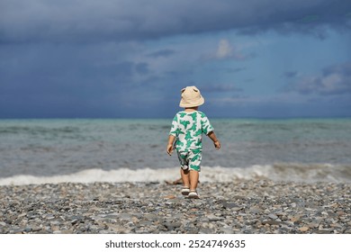 A toddler in a cute outfit and a sunhat walks along a rocky beach near the ocean as waves crash nearby. The scene captures the child's adventurous spirit and the natural beauty of the stormy  - Powered by Shutterstock