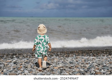 A toddler in a cute outfit and a sunhat walks along a rocky beach near the ocean as waves crash nearby. The scene captures the child's adventurous spirit and the natural beauty of the stormy  - Powered by Shutterstock