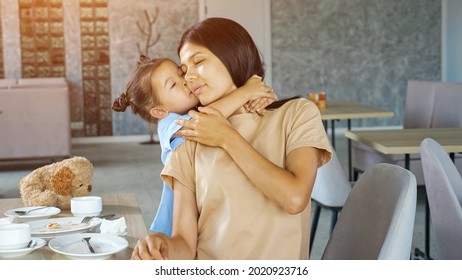 Toddler Daughter Hugs Mom. Little Child Girl Hugs Asian Mum Brunette With Smile Near Table With White Tea Cups In Local Restaurant Close View