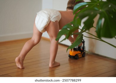 A toddler in a diaper crawling on a wooden floor, playing with a toy truck. The lush green plant adds a natural element, while the clean, light background offers space for text. The style is lifestyle - Powered by Shutterstock