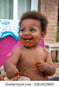 Toddler eats his dinner with no clothes on.  He has cute dimples in his cheeks as he laughs. sstkbabies. Happy smiling biracial  boy.
