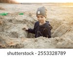 A toddler enjoys a day at the beach, digging in the sand with a green shovel. The background features a green net and a beautiful sunset, making the moment picturesque and tranquil