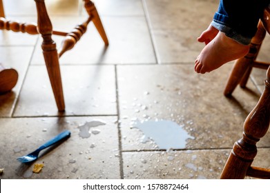 Toddler Foot Suspended Above Spilled Milk, Crumbs, And A Plastic Fork On A Brown Tile Floor 