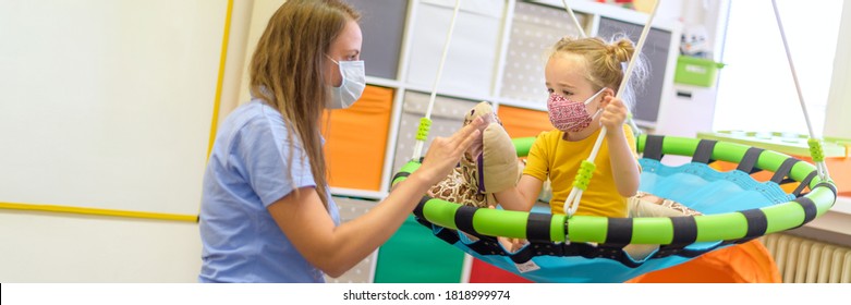 Toddler girl in child occupational therapy session doing playful exercises with her therapist during Covid - 19 pandemic, both wearing protective face masks. Web banner.