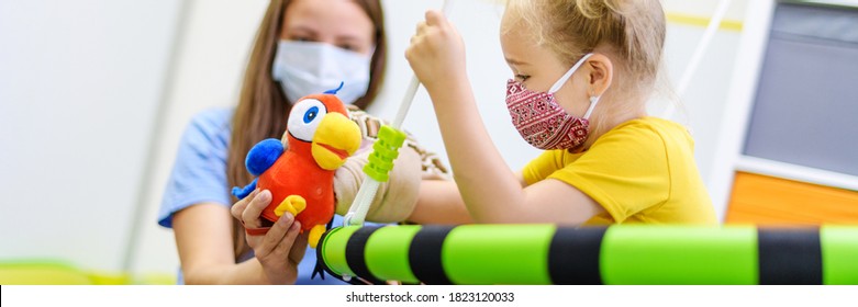 Toddler Girl In Child Occupational Therapy Session Doing Playful Exercises With Her Therapist During Covid - 19 Pandemic, Both Wearing Protective Face Masks. Web Banner.