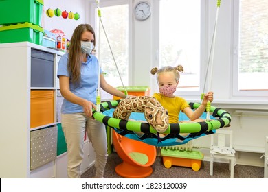 Toddler Girl In Child Occupational Therapy Session Doing Playful Exercises With Her Therapist During Covid - 19 Pandemic, Both Wearing Protective Face Masks.