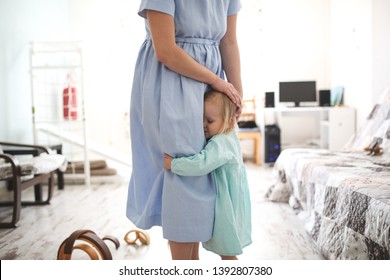 Toddler Girl Is Embarrassed And Hugs Mom By The Skirt, Hides Under The Skirt, The Concept Of Emotional Motherhood And Affection, Gentle Upbringing, Modest And Shy Child And Mother's Protection