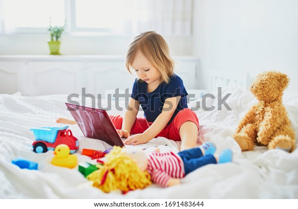 Toddler girl with laptop and toys in bed.
Kid using gadget to communicate with friends or kindergartners.
Education, distance learning or work from home with kids. Stay at
home entertainment