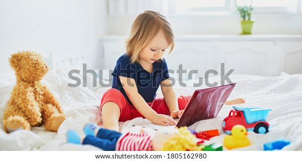 Toddler girl with laptop and toys in bed.\
Kid using gadget to communicate with friends or kindergartners.\
Education, distance learning or work from home with kids. Stay at\
home entertainment