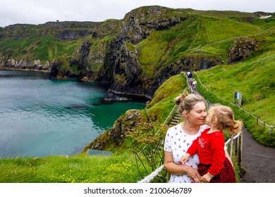 Toddler Girl And Mother On Carrick-a-Rede Rope Bridge, Famous Rope Bridge Near Ballintoy, Northern Ireland On Irish Coastline. Family Of Child And Woman On Bridge To Small Island On Cloudy Day.