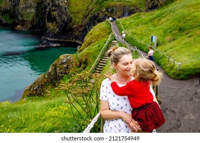 Toddler Girl And Mother On Carrick-a-Rede Rope Bridge, Famous Rope Bridge Near Ballintoy, Northern Ireland On Irish Coastline. Family Of Child And Woman On Bridge To Small Island On Cloudy Day.