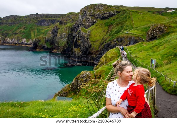 Toddler girl and mother on Carrick-a-Rede Rope\
Bridge, famous rope bridge near Ballintoy, Northern Ireland on\
Irish coastline. Family of child and woman on bridge to small\
island on cloudy day.