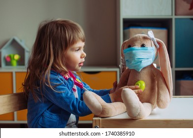 Toddler girl playing doctor using stethoscope toy with bunny soft toy in the face mask in the kids room. Pandemic COVID-19 concept.