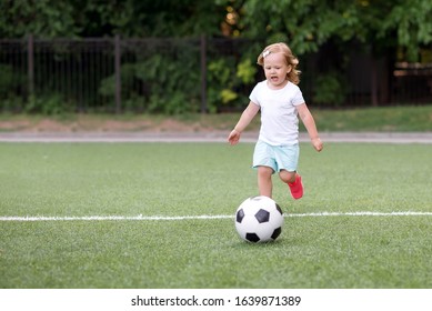 Toddler Girl Playing Football: Blonde Child In White Shirt And Pink Sneakers Running On Green Soccer Field To Kick Ball. Young Sportsman, Female Power And Active Childhood Concept. Copy Space