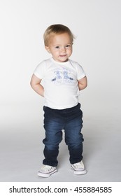 A Toddler Girl In White T-shirt And Blue Jeans Standing With Hands Behind Back On White Background