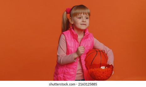 Toddler Happy Young Girl Kid Sportsman Basketball Fan In Pink Vest Holding And Eating Popcorn Doing Winner Gesture, Celebrating Victory Win Of Favourite Team Championship Isolated On Orange Background