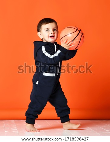Toddler infant baby kid child hold basketball ball happy smiling laughing ready for game play in dark blue hoodie costume on orange background