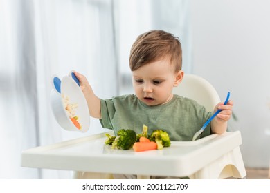 Toddler Kid Holding Spoon And Bowl Near Vegetables On High Chair