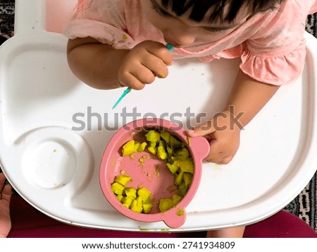 A toddler learning to eat independently using a spoon while sitting in a high chair. The child is enjoying freshly cut avocado pieces, practicing self-feeding skills during mealtime at home. The scene highlights early childhood development, healthy eating habits, and a nurturing family environment.