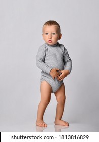 Toddler One-year-old Baby Boy In Grey One-piece Bodysuit With Long Sleeves Stands Looking At Camera Holding Hands At His Stomach. Happy Infancy And Babyhood Concept