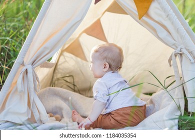 Toddler Playing In Cotton Teepee With Window, Decorated With Flags. One Year Cute Boy In A Teepee Outdoors.