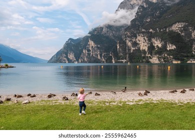 Toddler playing with group of ducks on pebble beach in lakeside town Nago-Torbole, Trentino, Italy. Lake Garda surrounded by steep rugged mountain peaks of Garda Prealps. Summer travel destination - Powered by Shutterstock