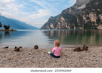 Toddler playing with group of ducks on pebble beach in lakeside town Nago-Torbole, Trentino, Italy. Lake Garda surrounded by steep rugged mountain peaks of Garda Prealps. Summer travel destination - Powered by Shutterstock