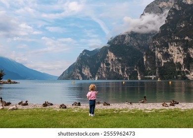 Toddler playing with group of ducks on pebble beach in lakeside town Nago-Torbole, Trentino, Italy. Lake Garda surrounded by steep rugged mountain peaks of Garda Prealps. Summer travel destination - Powered by Shutterstock