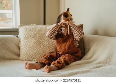 Toddler Playing With Wooden Camera And  Sitting On Cozy Couch By The Window