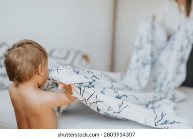 A toddler pulls a patterned duvet on a bed while his mother helps in the background. The child and mom share a quiet morning moment making the bed together.