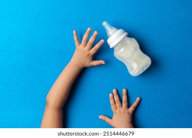 A toddler Reaching For milk feeding bottle, against blue back ground, top shot - Powered by Shutterstock