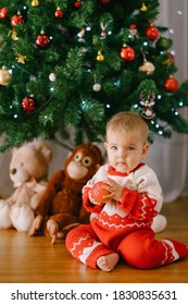 Toddler In A Red And White Christmas Onesie Is Holding An Apple In Front Of A Christmas Tree