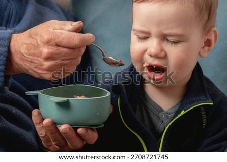 Toddler refusing to eat while grandfather tries to feed him with love and patience.