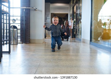 Toddler Running In The Shopping Center With His Mom On Background.