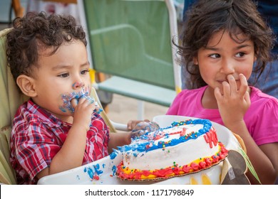 A Toddler Sets On A Baby High Chair Eating Cake With His Sister Next To Him Helping.