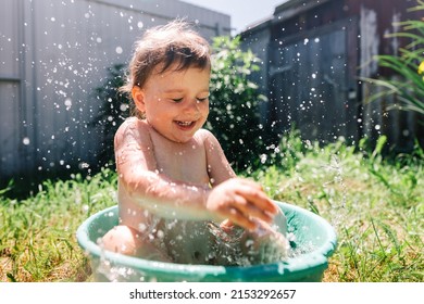 Toddler Sitting In Basin In Garden In Hot Summer Day Smiling Splashing Having Fun Not Looking At Camera. Horizontal Shot. Wide Angle View. Activities In Summertime And Carefree Childhood Concept.