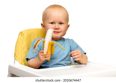 The toddler is sitting in a high chair holding a banana in his hand and smiling, laughing. Healthy nutrition of children with vitamins. Isolate the child of a boy or girl on a white background.