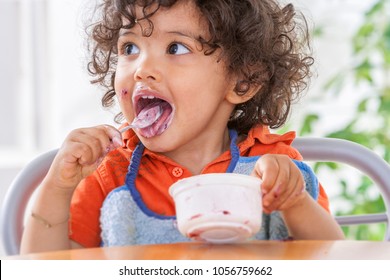 Toddler Sitting In Highchair And Eating Greek Yogurt. Baby Learning To Eat And Has Yogurt On Face.