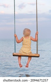 Toddler Sitting On Rope Swing Over The Water. Back View. Vertical Frame. Happy Childhood